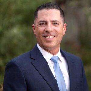 Photo of Corey Miller - a smiling man wearing a suit in front of a backdrop of trees in soft focus