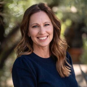 Photo of Cortney Critchfield - a smiling woman with long wavy hair in front of a backdrop of trees in soft focus