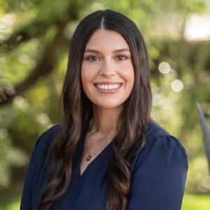 Photo of Sam Leedy - a smiling woman with long hair in front of a backdrop of trees in soft focus