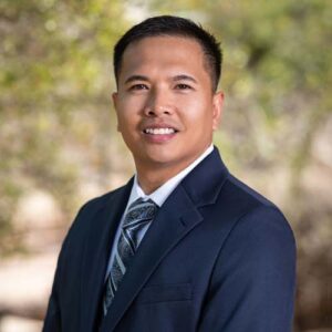 Photo of Brixon Tumaneng - a smiling man in a blue jacket and tie in front of trees