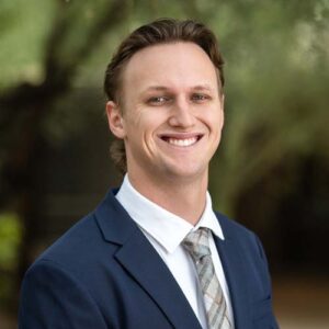 Photo of Luke Bell - a smiling man in a blue jacket and tie in front of trees